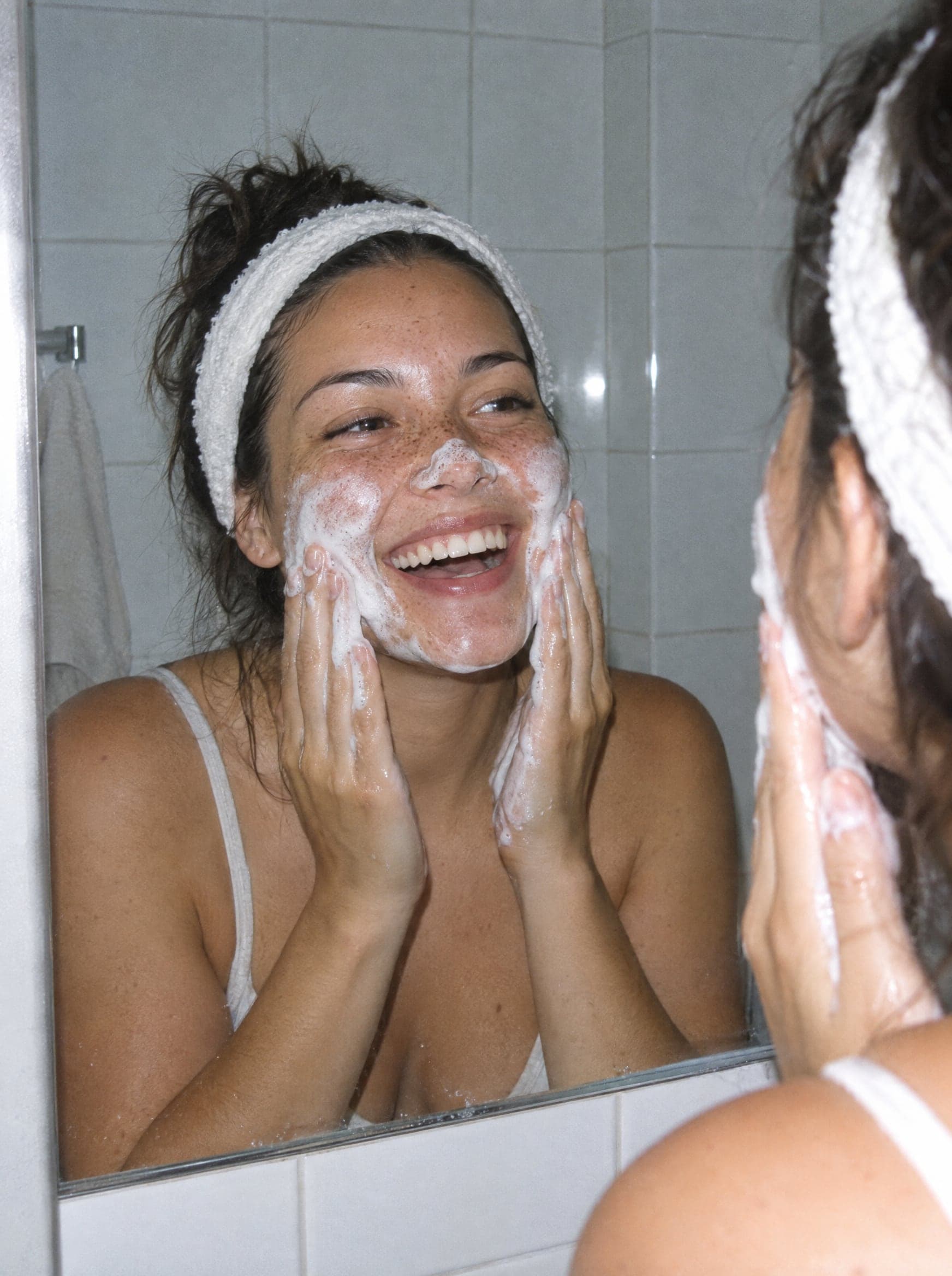 A woman in a headband laughing with cleanser foam on her face