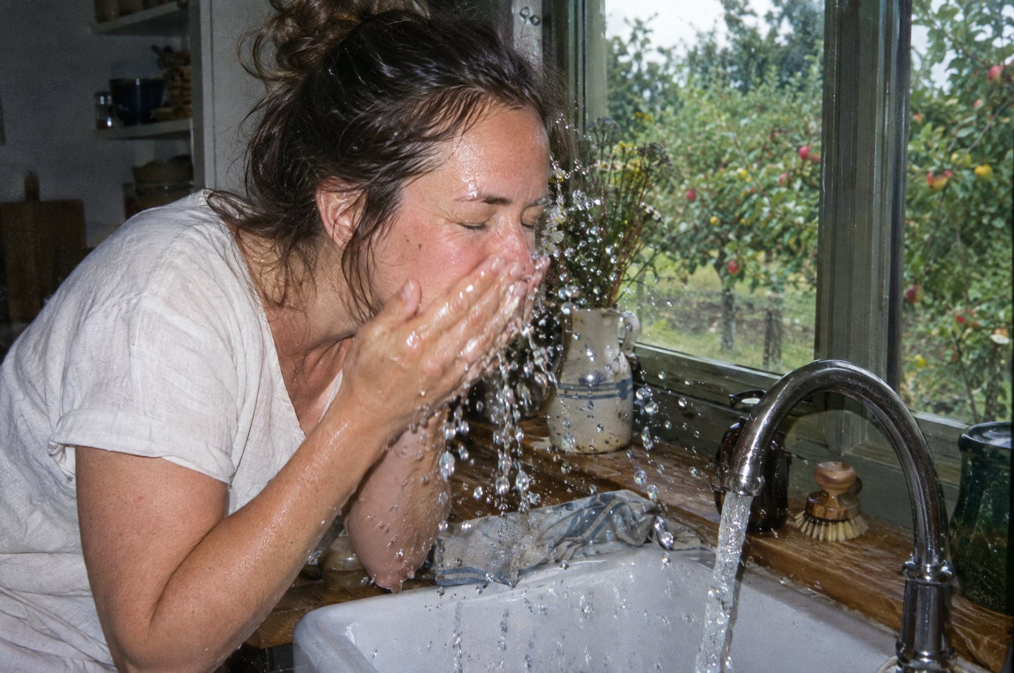A woman splashing water onto her face at a farmhouse kitchen sink