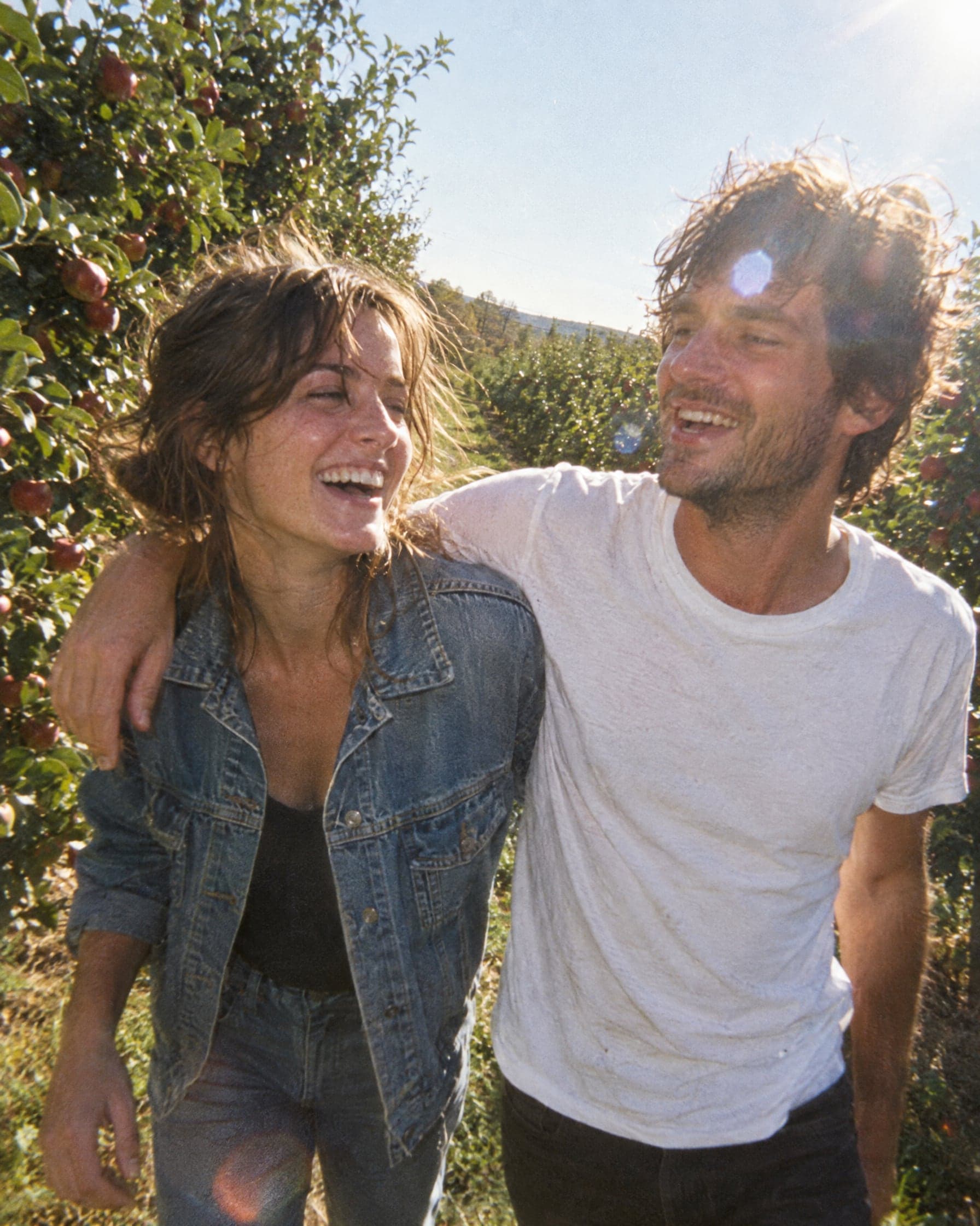 Two friends walking arm in arm between rows of apple trees in the sun